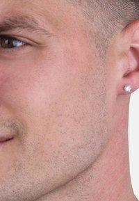 Silverfärgad studörhänge med en rund, texturerad design som bärs i örsnibben, tillsammans med en närbild av slät ansikts hud och kort hår.