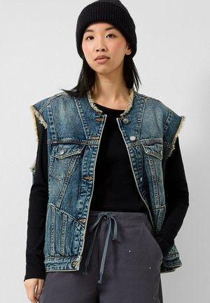 Jeune personne portant un bonnet noir, une chemise noire à manches longues, un gilet en jean effiloché et un pantalon gris avec cordon, les mains dans les poches.