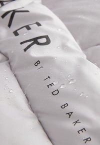 Biely vodotesný materiál má textúrovaný povrch s viditeľnými kvapkami. Čierny text „BAKER od TED BAKER“ je výrazne znázornený.