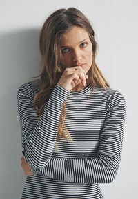 Gestreiftes, langärmeliges Oberteil in Schwarz und Weiß; figurbetontes Design mit gerippter Textur. Model posiert mit dem Kinn auf der Hand und präsentiert das Kleidungsstück.