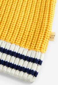 Gelbes gestricktes Gewebe mit einer strukturierten Oberfläche, das am unteren Rand ein gestreiftes Muster in Weiß und Marineblau aufweist. Kleines Etikett sichtbar.