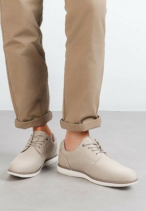 Baskets décontractées beiges avec lacets portées avec un pantalon brun clair retroussé sur un sol gris.