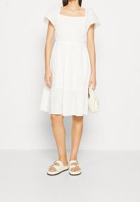 Robe blanche à plusieurs niveaux avec des manches bouffantes et un décolleté carré. Tissu lisse avec une texture subtile. Le mannequin porte des sandales à glisser beiges et tient une pochette.