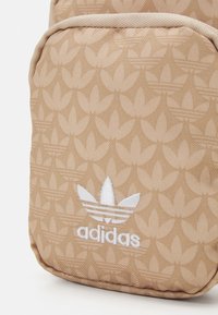 Béžový batoh s texturovaným květinovým vzorem, lemovaný kontrastní látkou. Na přední straně je bílý vyšívaný motiv adidas.