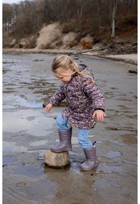 Bambino con giacca imbottita viola a fiori in piedi su una roccia, indossa stivali di gomma grigi e jeans azzurri vicino a una riva fangosa.