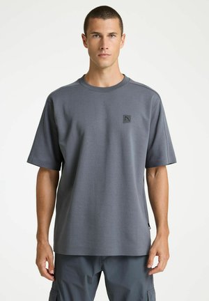 Jeune homme aux cheveux courts portant un t-shirt gris uni surdimensionné et un pantalon gris foncé assorti, debout devant un fond blanc.