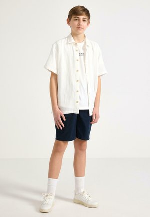 Teenagedreng stående iført hvid åben kortærmet skjorte over hvid T-shirt, marineblå shorts, hvide sokker og hvide sneakers mod en ensfarvet baggrund.