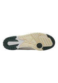 Suela de zapatilla New Balance en negro y crema, con patrones texturizados, surcos y un acento circular. El material parece ser de goma para proporcionar tracción.