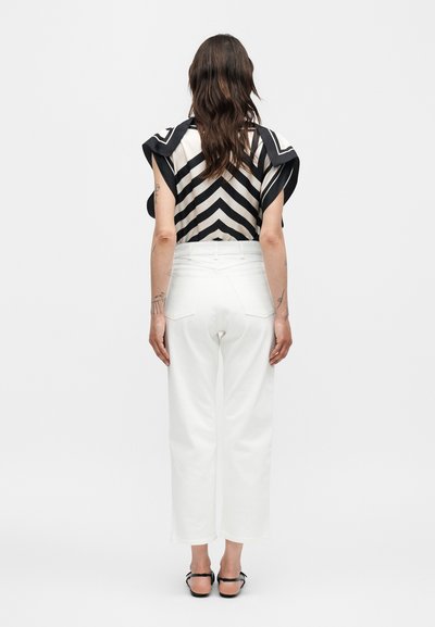 Γυναίκα με μακριά σκούρα μαλλιά, φοράει τοπ με ασπρόμαυρο σχέδιο ζιγκ-ζαγκ και άσπρο, φαρδύ, ψηλόμεσο παντελόνι, με την πλάτη προς τα εμάς.