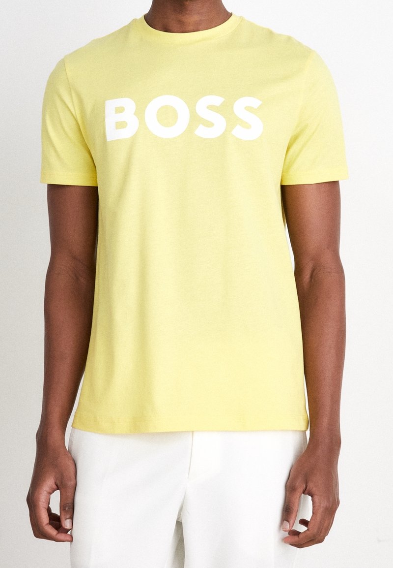Man wearing a yellow short-sleeve t-shirt with "BOSS" in white letters, paired with white pants, against a plain background.
