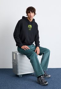 Černá mikina s kapucí a zeleným potiskem, ve spojení s tyrkysovými kalhotami. Model sedí na stříbrném kovovém boxu. Šedé tenisky doplňují outfit.