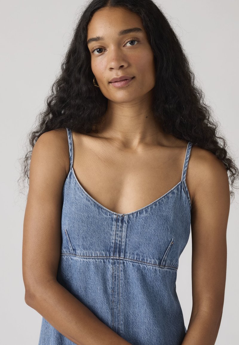 Frau mit langen lockigen Haaren, die ein blaues ärmelloses Jeanskleid trägt und vor einem schlichten hellen Hintergrund steht, schaut in die Kamera.