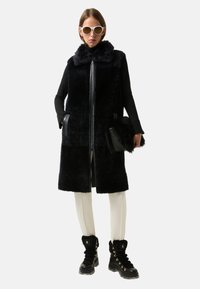 Czarna, sztuczna futrzana kamizelka bez rękawów z futrzanym kołnierzem, w połączeniu z białymi spodniami i czarnymi, sznurowanymi botkami z złotymi akcentami.