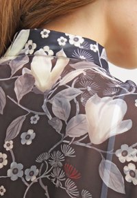 Durchsichtige schwarze Bluse mit floralen Mustern in sanften Farben, die Magnolien und kleine Blumen hervorheben. Mit Kragen und dezenten Texturen.