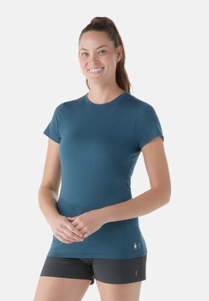 Femme souriante avec une queue de cheval, portant un T-shirt ajusté turquoise et un short noir, debout les mains jointes devant sur un fond blanc.
