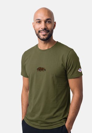 Lächelnder Mann mit Bart, trägt ein olivgrünes T-Shirt mit einer kleinen Bärengrafik auf der Brust und einem Aufnäher am linken Ärmel, Hände in den Taschen.