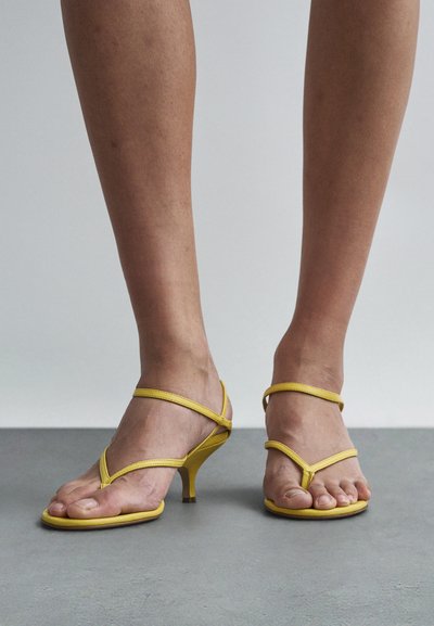 Nahé nohy v žlutých sandálech na podpatku s pásky, stojící na šedé podlaze před jednobarevným světlým pozadím.