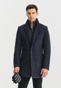 Czarny wełniany płaszcz o eleganckim kroju, z kołnierzem typu lapel, z przodu zapinany na guziki, z pikowaną warstwą wewnętrzną, w połączeniu z czarnymi skórzanymi rękawiczkami.