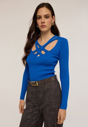 Top a maniche lunghe a coste di un vivace blu, caratterizzato da un design del collo incrociato unico. Abbinato a pantaloni a quadri scuri e una cintura marrone.