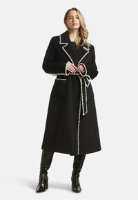 Black wool coat with contrasting white trim, large collar, two front pockets, and a matching belt; mid-length design, soft texture.