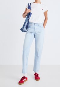 Camiseta blanca de manga corta con un pequeño logo rojo, jeans rectos de mezclilla azul claro, zapatillas de ante rojas con rayas blancas, llevando una bolsa de mezclilla.