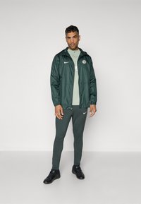 Green nylon jacket with a hood and Nike logo, paired with a light green shirt and matching fitted pants. Black athletic shoes.