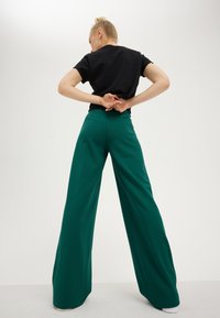 Personne aux cheveux blonds attachés en chignon, portant un t-shirt noir et un pantalon large vert, debout les mains jointes derrière le dos sur un fond uni.