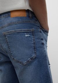 Siniset farkut, joissa on kulunut muotoilu, repaleinen tasku, ruskea nahkavaateetäge ja perinteinen viisi-taskun tyyli.