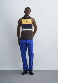 Homme debout, portant un débardeur à blocs de couleurs avec des rayures jaunes, bleues, marron et blanches, un pantalon bleu, des chaussures noires et un collier avec pendentif.