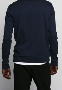 Felpa blu navy con maniche lunghe, polsini e orlo a costine. Realizzata in morbido tessuto testurizzato, indossata sopra una maglietta bianca. Design minimale.