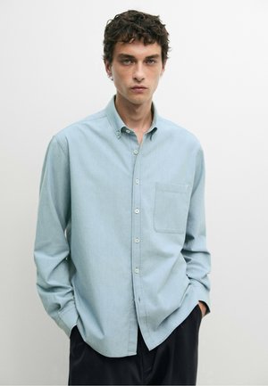 Giovane uomo con capelli scuri e ricci che indossa una camicia azzurra con bottoni e taschino sul petto e pantaloni neri, in piedi davanti a uno sfondo semplice.