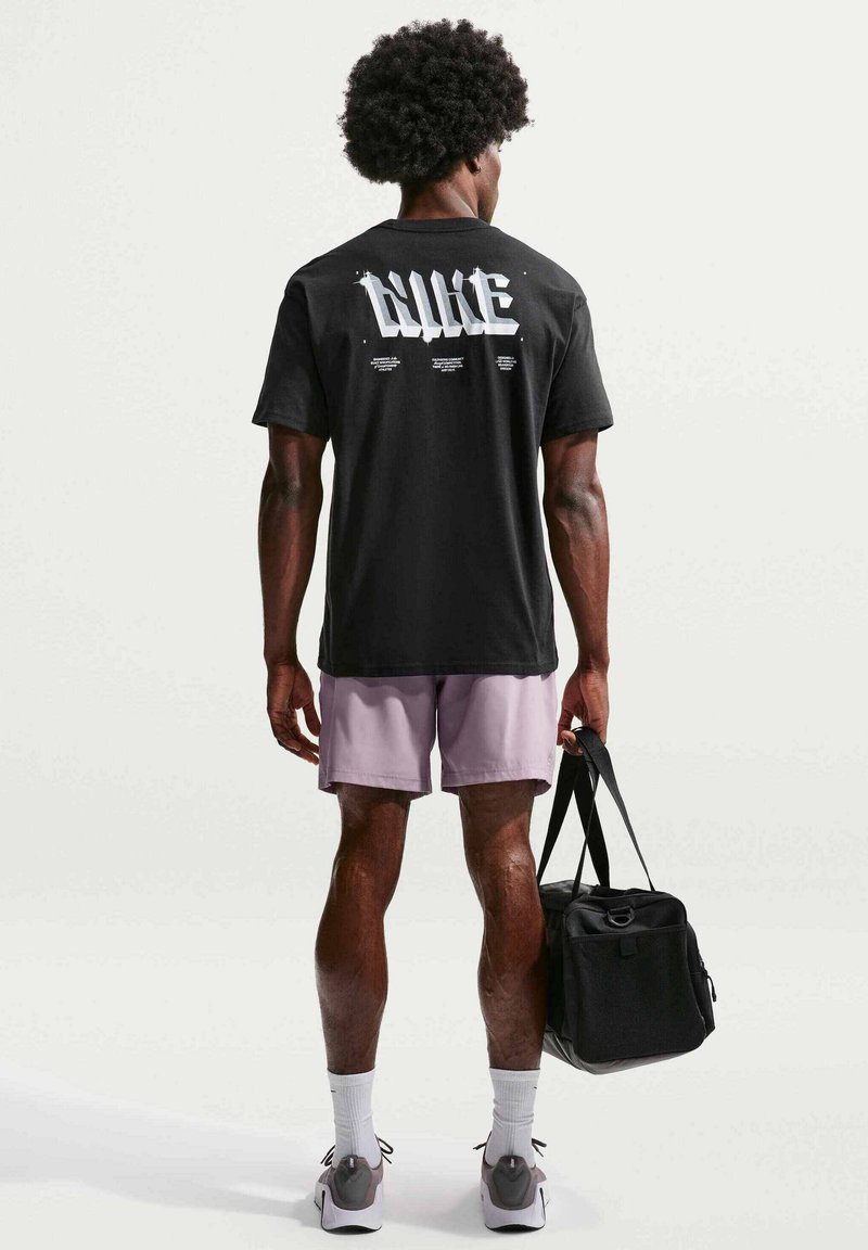 Homme avec coiffure afro portant un t-shirt Nike noir et un short violet, tenant un sac de sport noir, debout devant un fond uni.