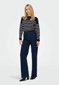 Top a righe navy e bianche con maniche lunghe e inserti a forma di cuore rossa, abbinato a jeans blu scuri a vita alta e stivali neri con tacco a blocco.