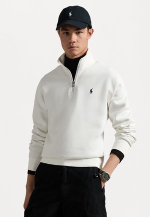 Felpa bianca con zip e logo nero, abbinata a un cappellino nero e pantaloni scuri. L'orologio al polso aggiunge un tocco di dettaglio all'outfit.
