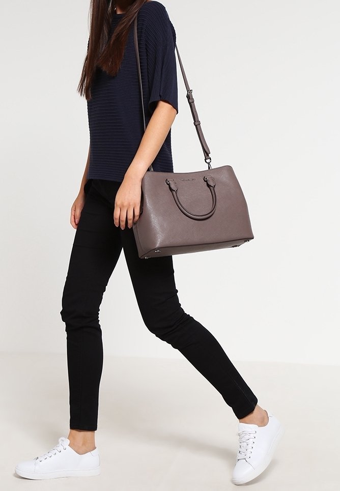 Femme en haut bleu marine, pantalon noir et baskets blanches portant un grand sac à main taupe à bandoulière, marchant sur un fond uni.