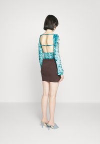 Top de veludo turquesa de mangas longas com recorte nas costas e detalhe de amarração, combinado com uma saia mini justa castanha e mules azuis de salto.