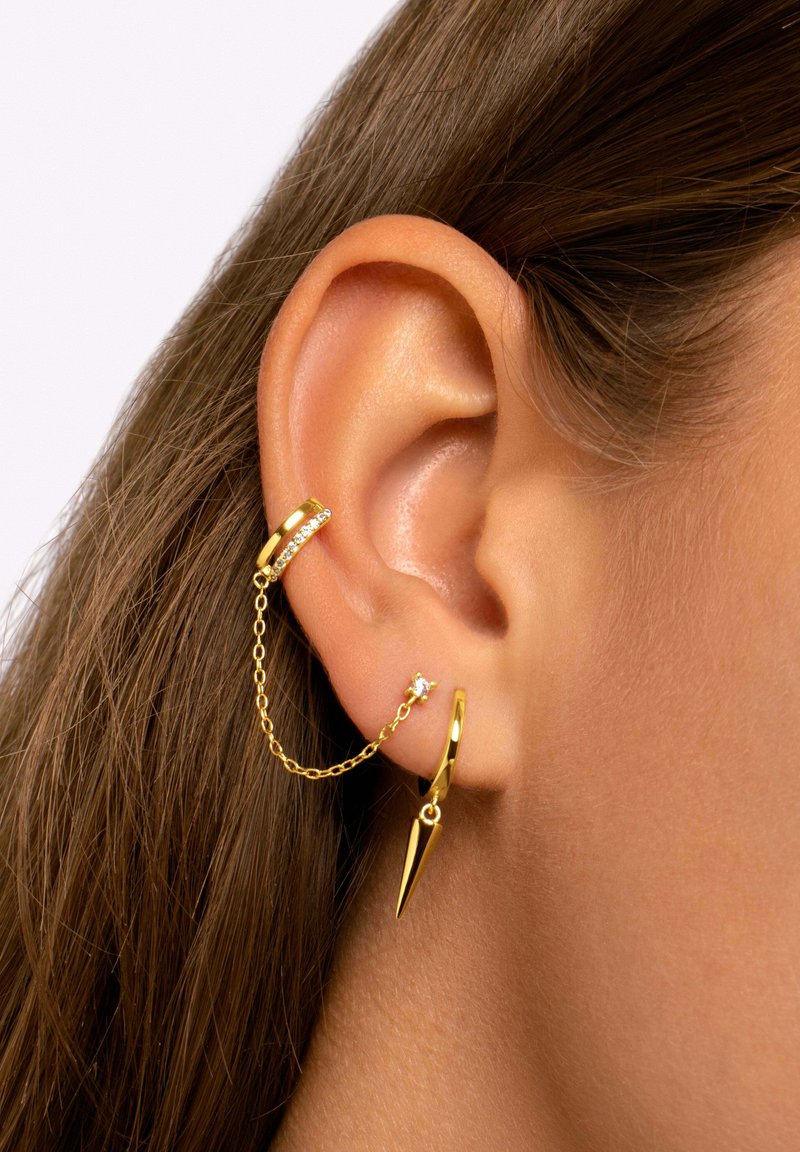 Orecchini d'oro con una catena che collega un cerchio con un accento in cristallo e un ciondolo a punta, che mostrano un design elegante e moderno.
