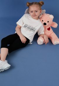 Jeune fille aux cheveux blonds coiffés en chignons, portant un t-shirt blanc et des leggings noirs, assise sur un sol bleu, tenant un ours en peluche rose avec un cœur.