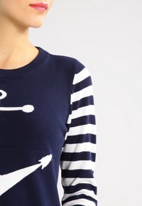 Kvinna som bär en marinblå tröja med vita nautiska pilar på framsidan och vita och marinblå horisontella randiga ärmar.