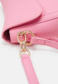 Gros plan d’un sac à main rose texturé avec fermoir en métal doré reliant la sangle au corps du sac sur fond blanc.