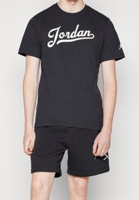 Czarny bawełniany t-shirt z białym logo "Jordan" w formie napisu, noszony z dopasowanymi czarnymi szortami, o gładkiej strukturze i swobodnym kroju.