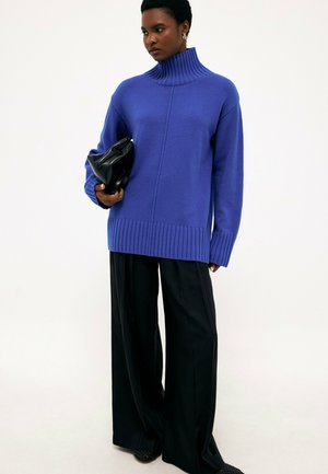 Donna che indossa un maglione dolcevita blu e pantaloni neri a gamba larga, tiene in mano una pochette nera piegata, in piedi su uno sfondo chiaro.