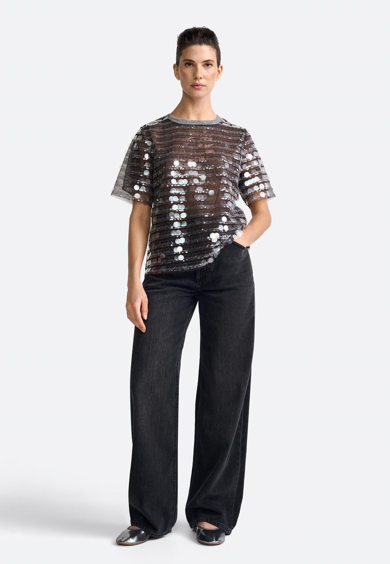 Gennemsigtig sort top udsmykket med metallic pailletter i et stribet mønster, parret med løse sorte jeans og enkle flade sko.