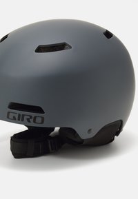 Casco grigio opaco con fori di ventilazione, cintura regolabile e branding nero sul lato. Superficie liscia con forma aerodinamica.