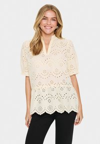 Hellbeige Bluse mit Lochmuster, gewelltem Saum und einem Schlüsselaugen-Ausschnitt. Kurze Puffärmel verleihen dem Design Volumen.