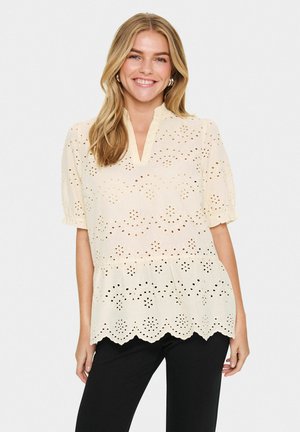 Blouse beige clair avec motifs en œillet, ourlet en dentelle et décolleté en clé. Les manches bouffantes courtes ajoutent du volume au design.