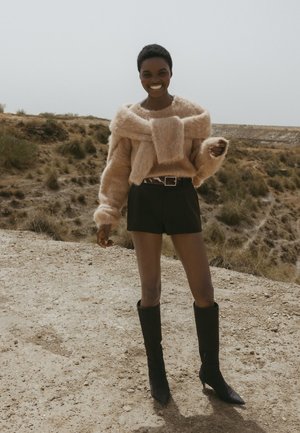 Luftig beige sweater med en stor sløjfedetalje, parret med sorte højtaljede shorts og knæhøje sorte støvler på en struktureret leroverflade.