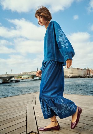 Denimový outfit s dlouhými rukávy a volánkovou sukní, zdobený květinovou výšivkou. Celkový vzhled doplňují ploché, lesklé vínové boty.
