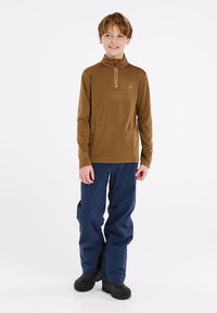 Bruin langmouw top met een halve rits, met een geaccentueerde textuur op de borst; gecombineerd met blauwe waterdichte broek en zwarte laarzen.