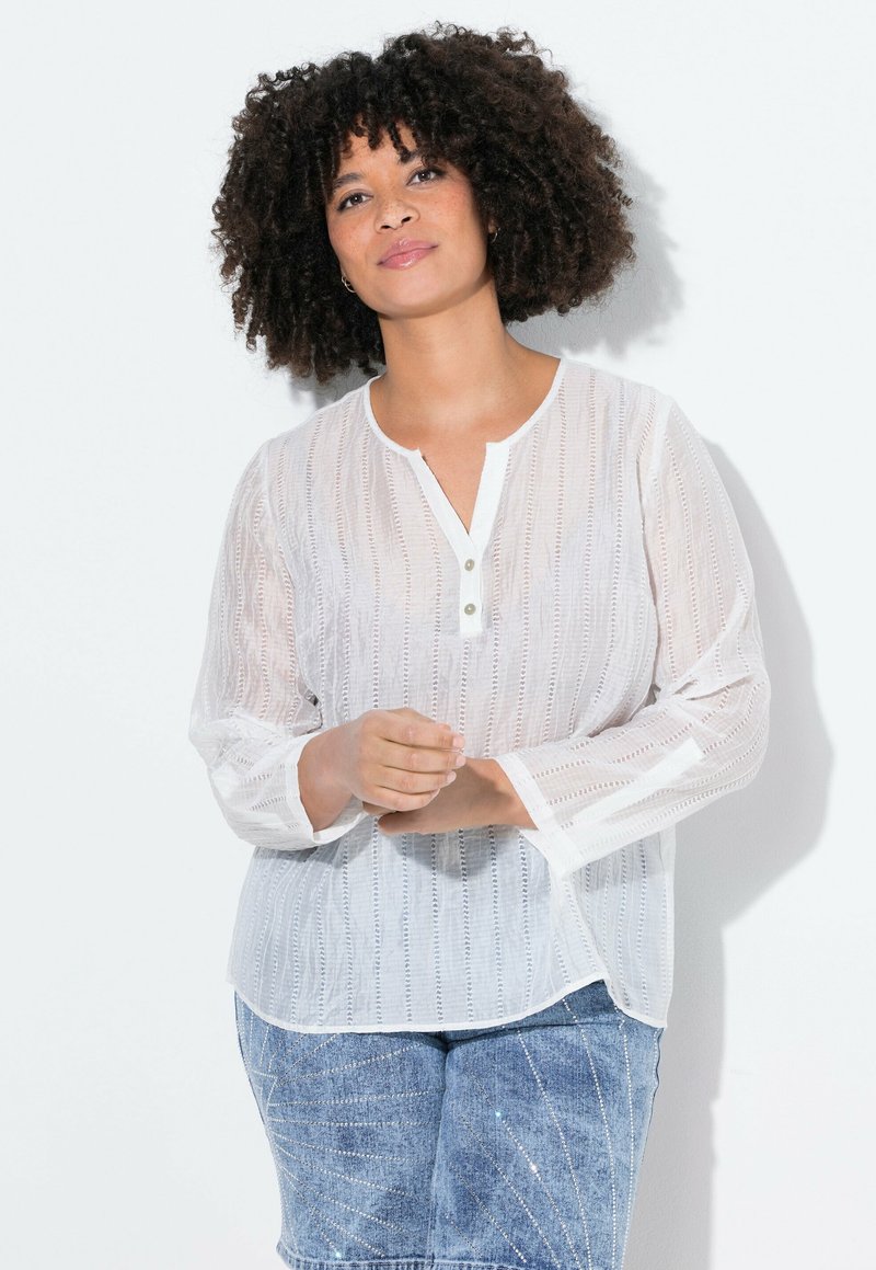 Blusa blanca de manga larga, hecha de un tejido liviano y texturizado, con una abertura de botones y sutiles rayas verticales. Combinada con jeans azules.